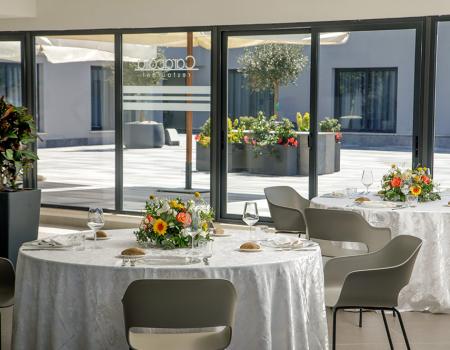 Sala elegante con tavoli rotondi, fiori colorati e luce solare intensa.