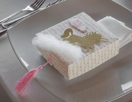 Elegante scatola con unicorno dorato, soffice cotone e nappina rosa su piatto raffinato.
